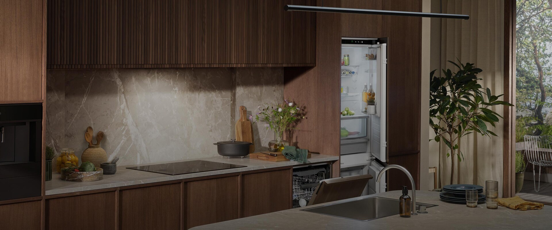 Integreret kølefryseskab og opvaskemaskine i et stilrent køkken med mørke træpaneler og bordplade i marmor.